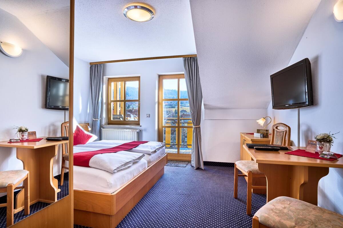 A hotel room with a bed, desk, chair, television, and a view of the city through large windows.