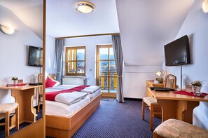 A hotel room with a bed, desk, chair, television, and a view of the city through large windows. - Arber-Hotel am Rothbach (Bodenmais)
