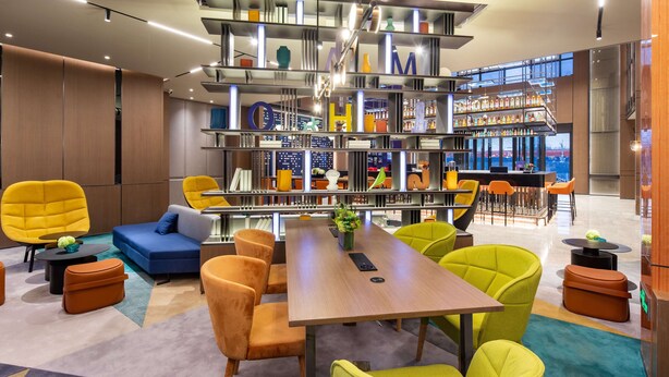 A modern lounge area with a central wooden table, vibrant yellow and green chairs, a blue sofa, and a bookshelf with various items. - Hampton by Hilton Beijing Wuzi Xueyuan Road (Tongzhou)