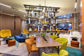 A modern lounge area with a central wooden table, vibrant yellow and green chairs, a blue sofa, and a bookshelf with various items. - Hampton by Hilton Beijing Wuzi Xueyuan Road (Tongzhou)