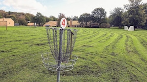 Una canasta de disc golf con un '4' rojo, rodeada por una cerca de malla metálica, situada en un campo cubierto de hierba con casas al fondo.