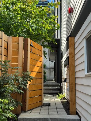 Un étroit sentier menant à l’entrée d’un bâtiment, bordé d’une clôture en bois et d’un arbre.