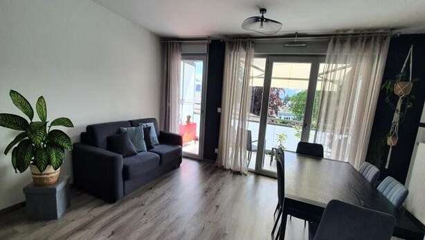 A modern living room with a sofa, dining table, chairs, and a potted plant. - Residence Le Clos Des Merisiers (Aix-les-Bains)