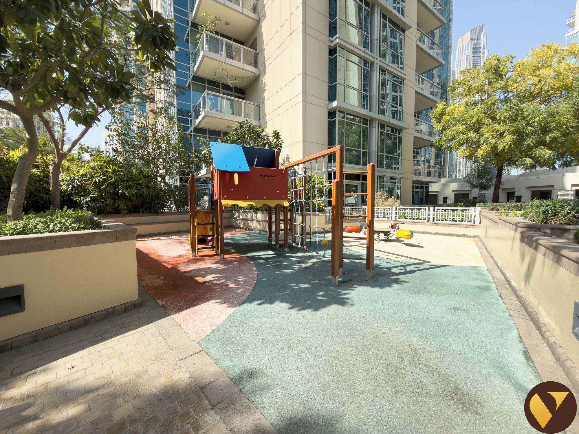 A playground with a climbing structure and swings, surrounded by a building with large windows and a paved area.