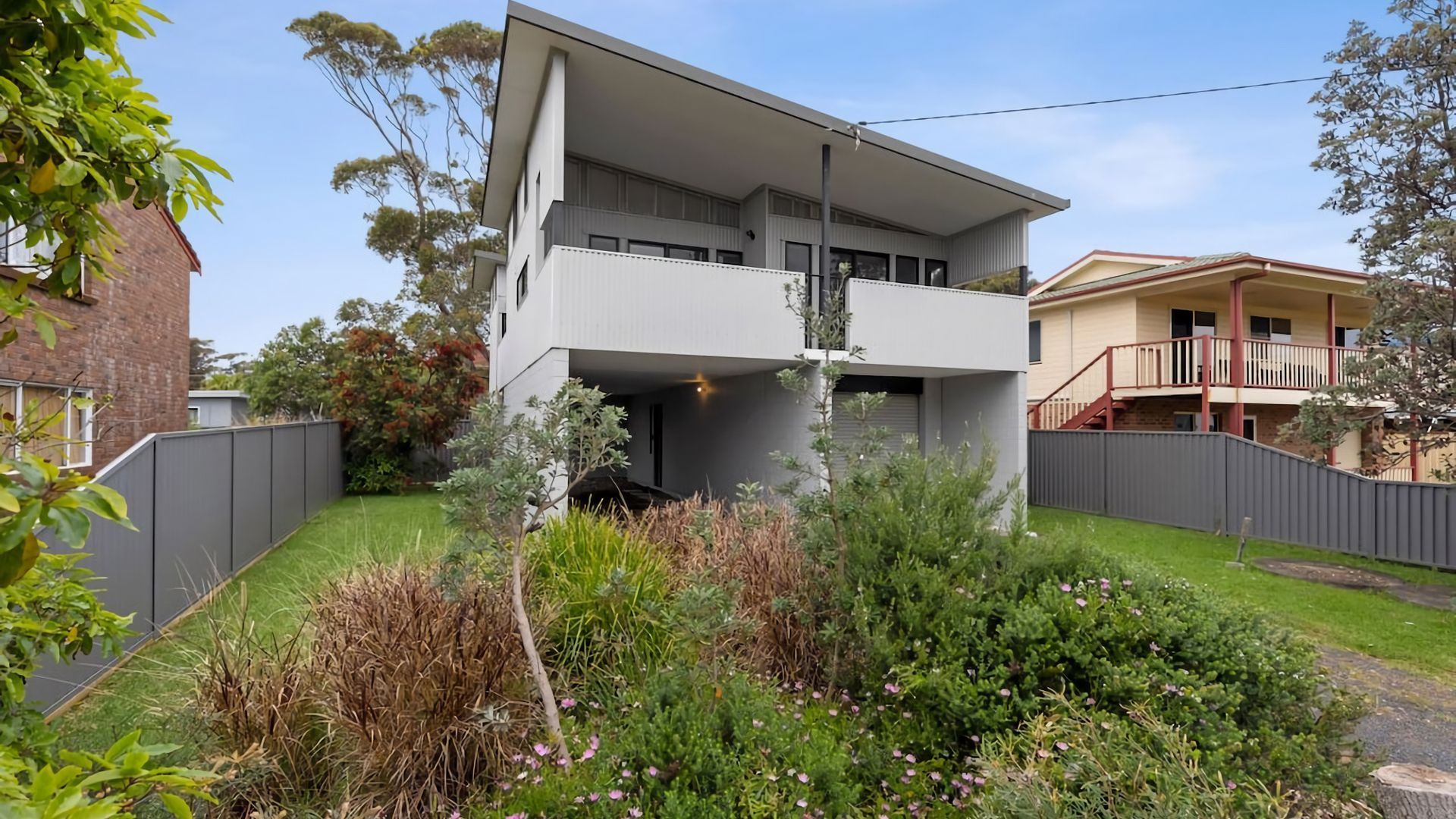A two-story residential building with a flat roof, a balcony on the upper floor, and a fenced backyard with various plants and shrubs.