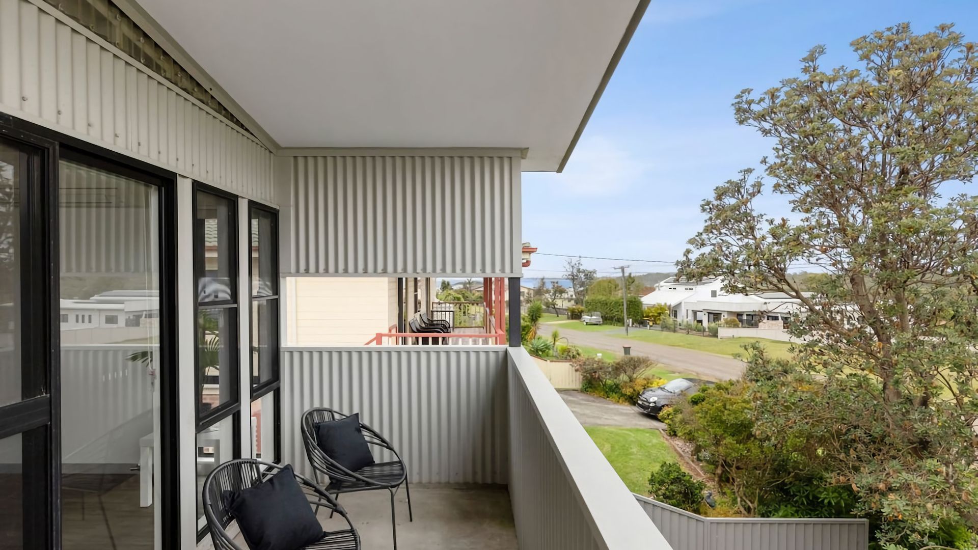 Un balcon avec deux chaises et une table, donnant sur une rue résidentielle bordée de maisons et d’un arbre.