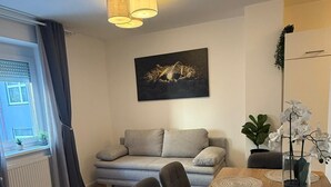 A modern living room with a sofa, a dining table with chairs, a large window with curtains, a ceiling light fixture, and a wall-mounted painting. - Central Area 1BR Apartment With Balcony (Vienna)