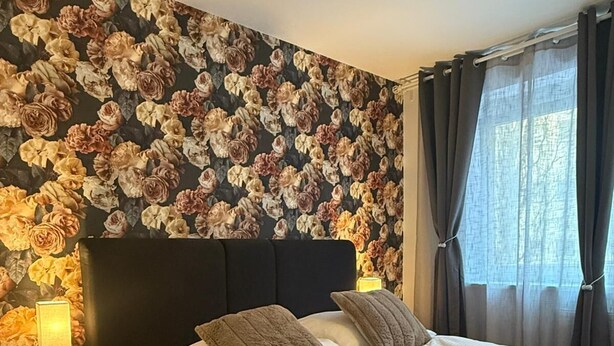 A neatly made bed with a dark headboard, a floral wallpaper, and a bedside lamp. - Central Area 1BR Apartment With Balcony (Vienna)