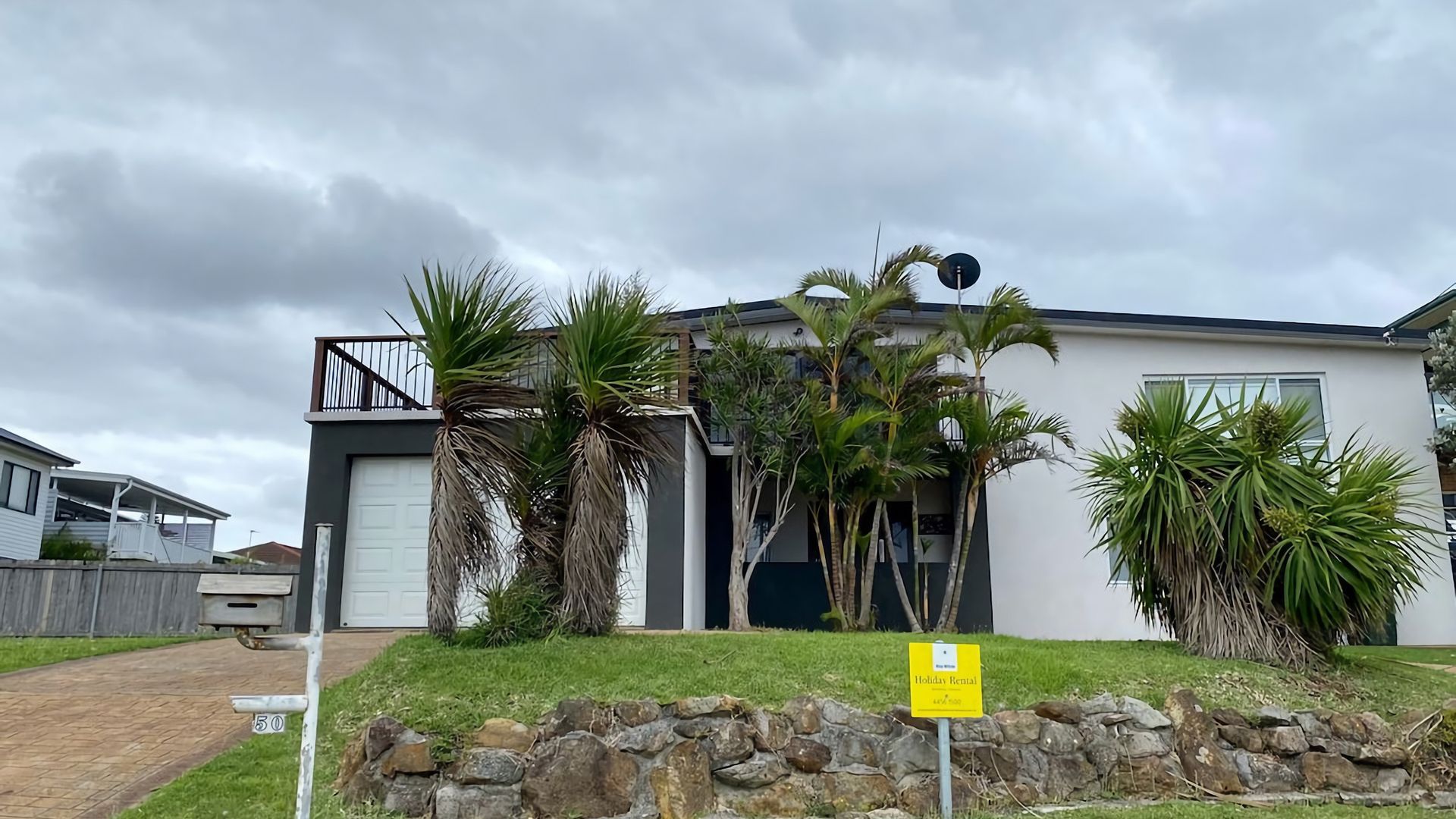 A modern two-story house with a garage, surrounded by a stone wall and palm trees.