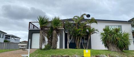 A modern two-story house with a garage, surrounded by a stone wall and palm trees.