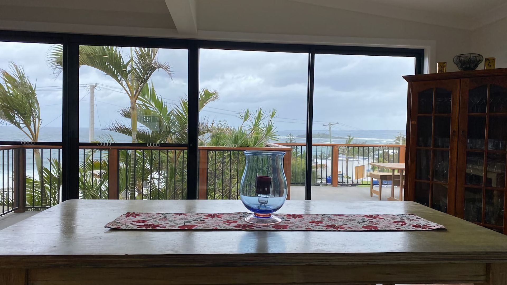 A balcony with a view of palm trees and the ocean, a glass vase on a table, and a wooden cabinet.