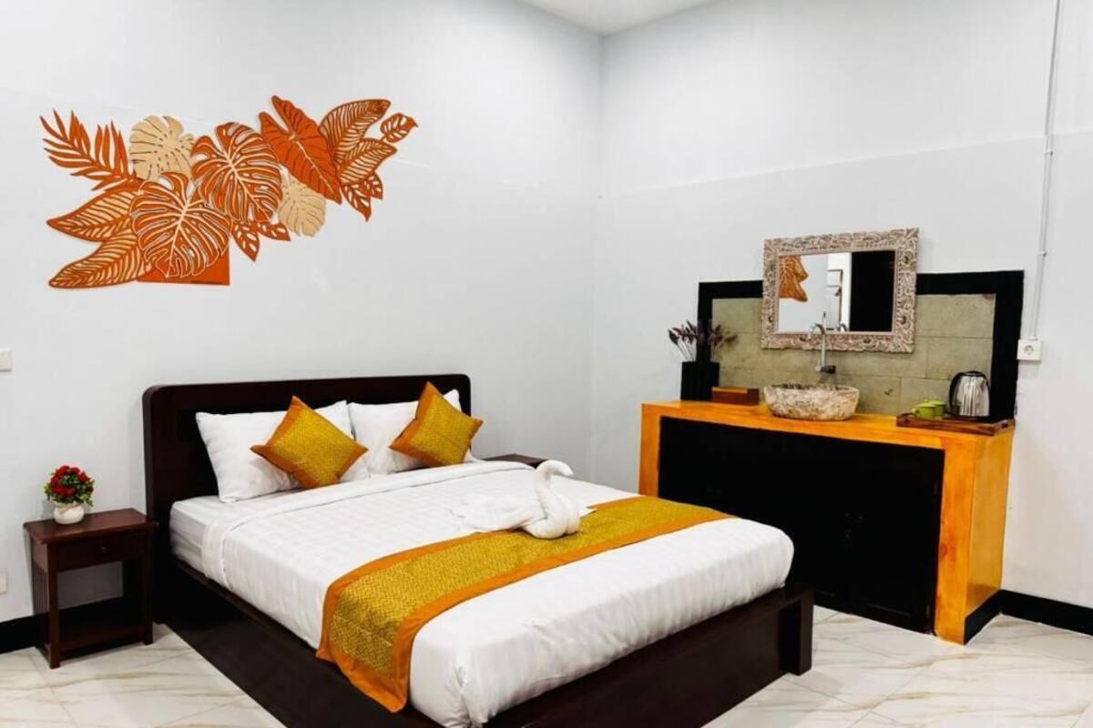 Una cama bien hecha con sábanas blancas y un detalle amarillo, una mesita de noche de madera con un jarrón de flores, un espejo sobre una consola de madera, y una pared con un adhesivo de hoja.
