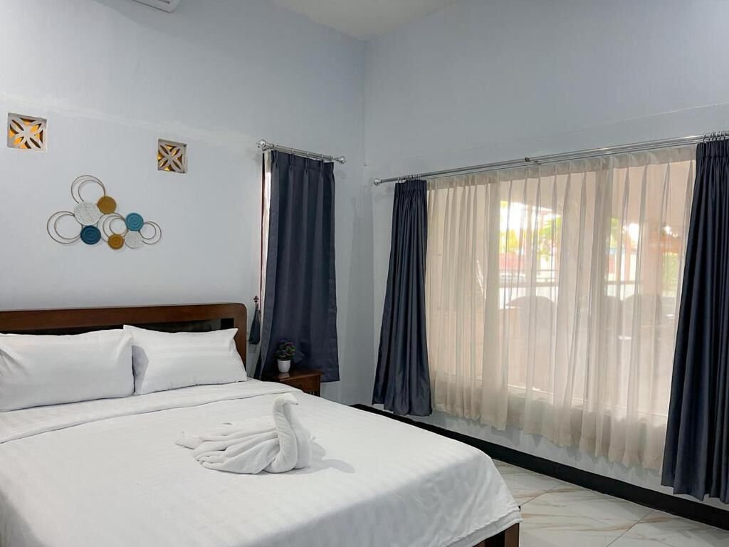 Una habitación de hotel con una cama bien hecha, una mesita de noche pequeña con una planta y una ventana con cortinas traslúcidas.