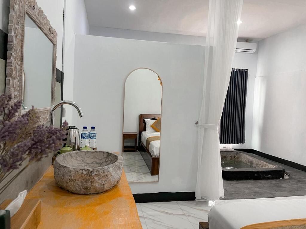 A modern hotel room with a marble sink, a large mirror, a bed with a wooden headboard, a small bedside table with toiletries, and a private outdoor shower area.