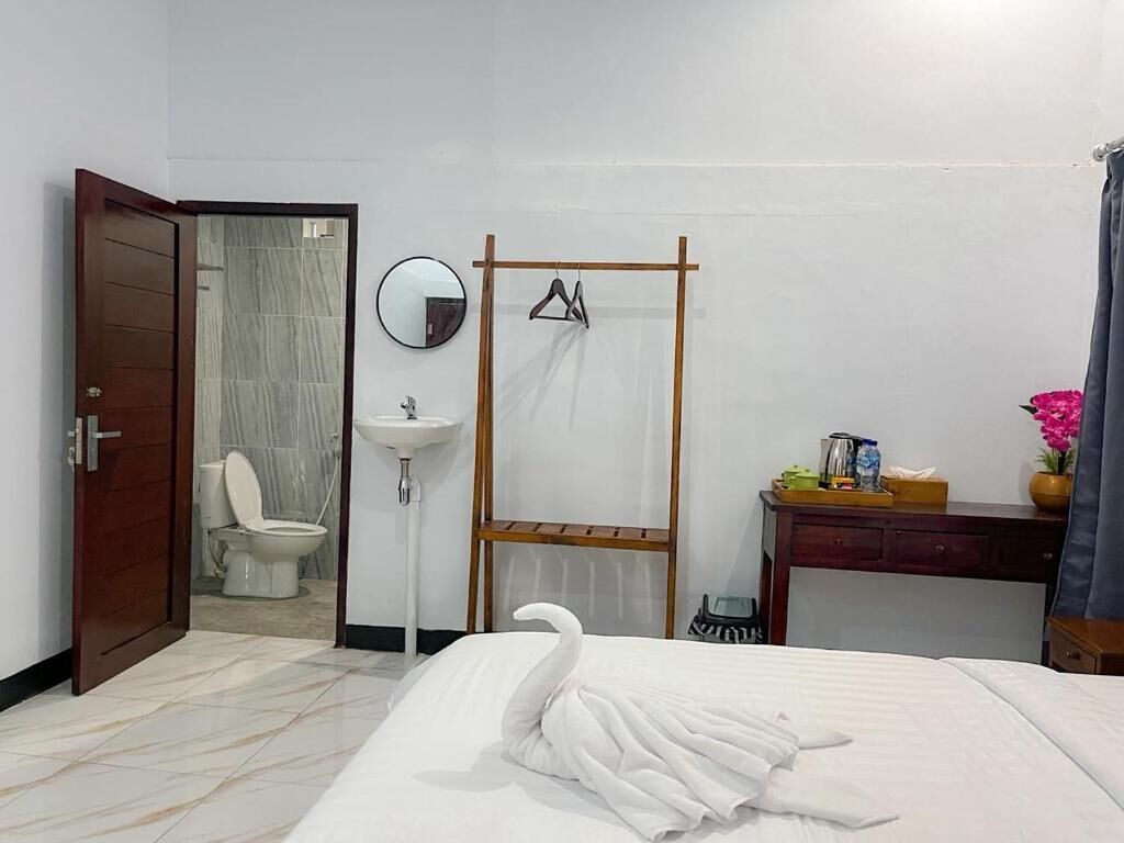 A hotel room with a bed, a wooden wardrobe, a bedside table with a lamp, a mirror, and a bathroom visible through an open door.