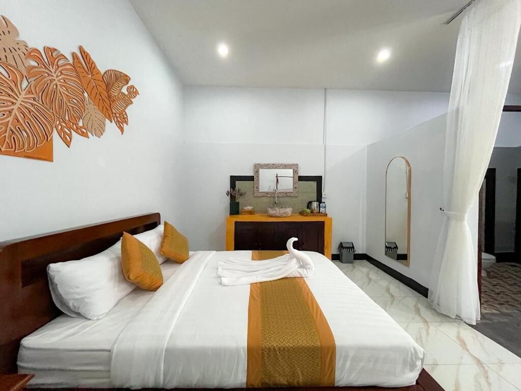 A hotel room with a neatly made bed, a small bedside table with a mirror, a sink, and a shower area.