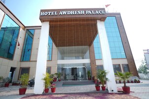 A entrada do Hotel Awdhesh Palace, com fachada moderna, grandes janelas de vidro e plantas em vasos à frente.