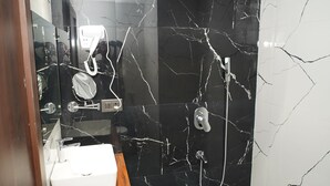 A bathroom with a black marble wall, a wooden vanity, a white sink, a shower with a handheld showerhead, and a small trash bin. - Avdhesh Palace (Tarabganj)
