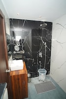 Casa de banho com parede de mármore preto, toucador de madeira, lavatório branco, chuveiro com chuveiro manual e um pequeno contentor de lixo.