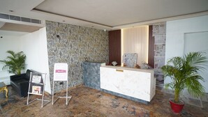A reception area with a marble counter, a reception desk, a wall with a textured pattern, a potted plant, and a black sofa. - Avdhesh Palace (Tarabganj)