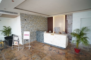 Uma área de recepção com balcão de mármore, mesa de atendimento, parede com textura, planta em vaso e sofá preto.