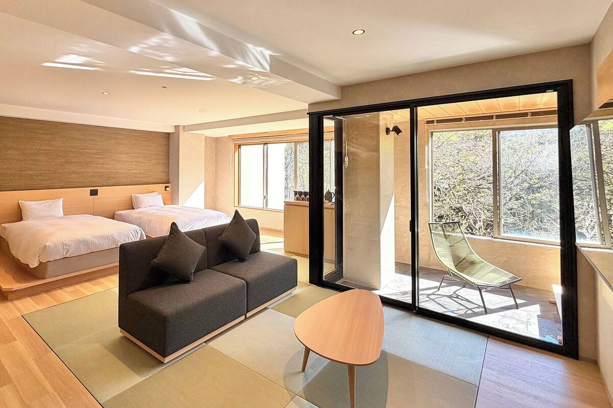 A modern hotel room with two beds, a sofa, a small table, and a sliding glass door leading to a balcony.