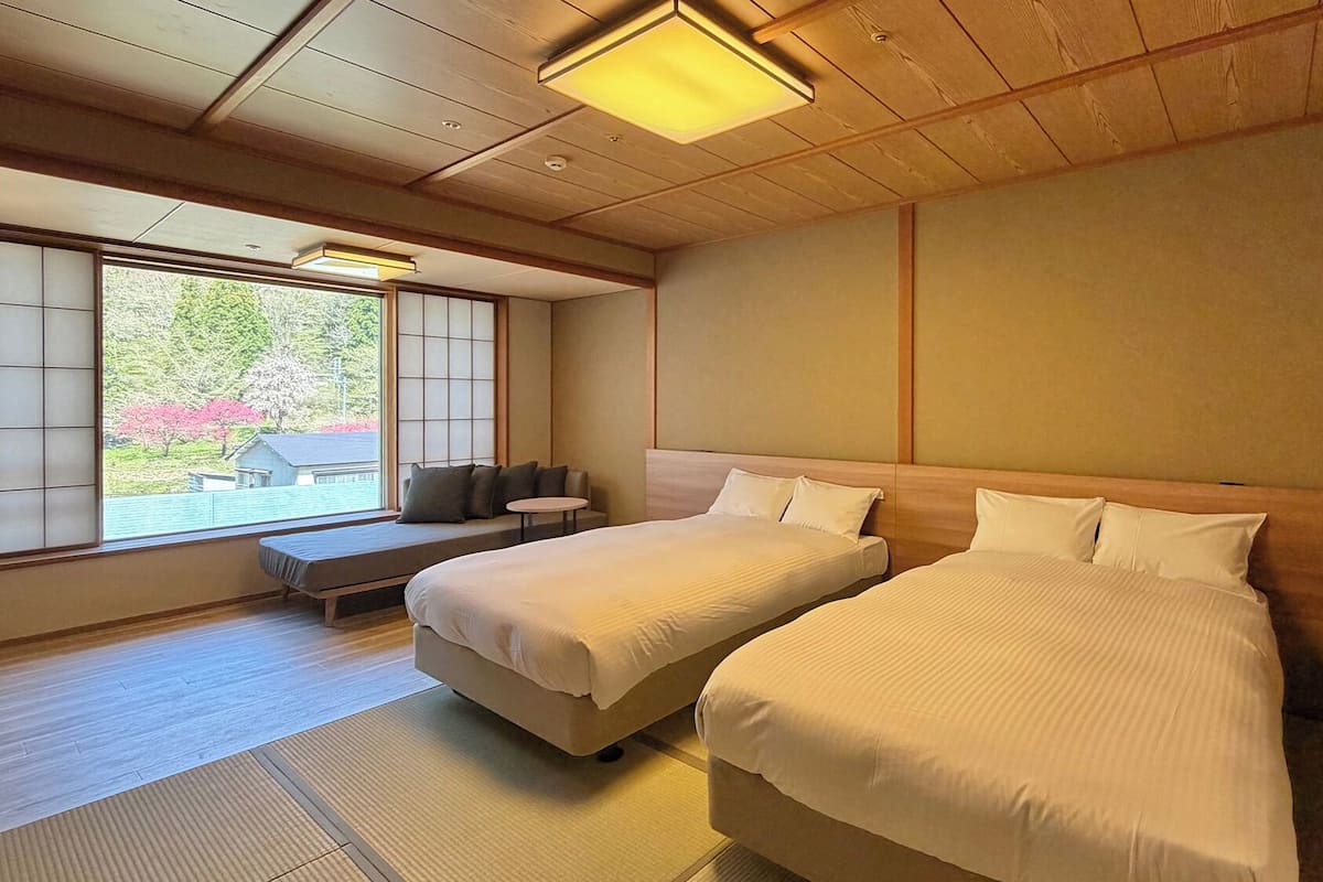 A room with two beds, a bench, and a view of trees and a house outside.