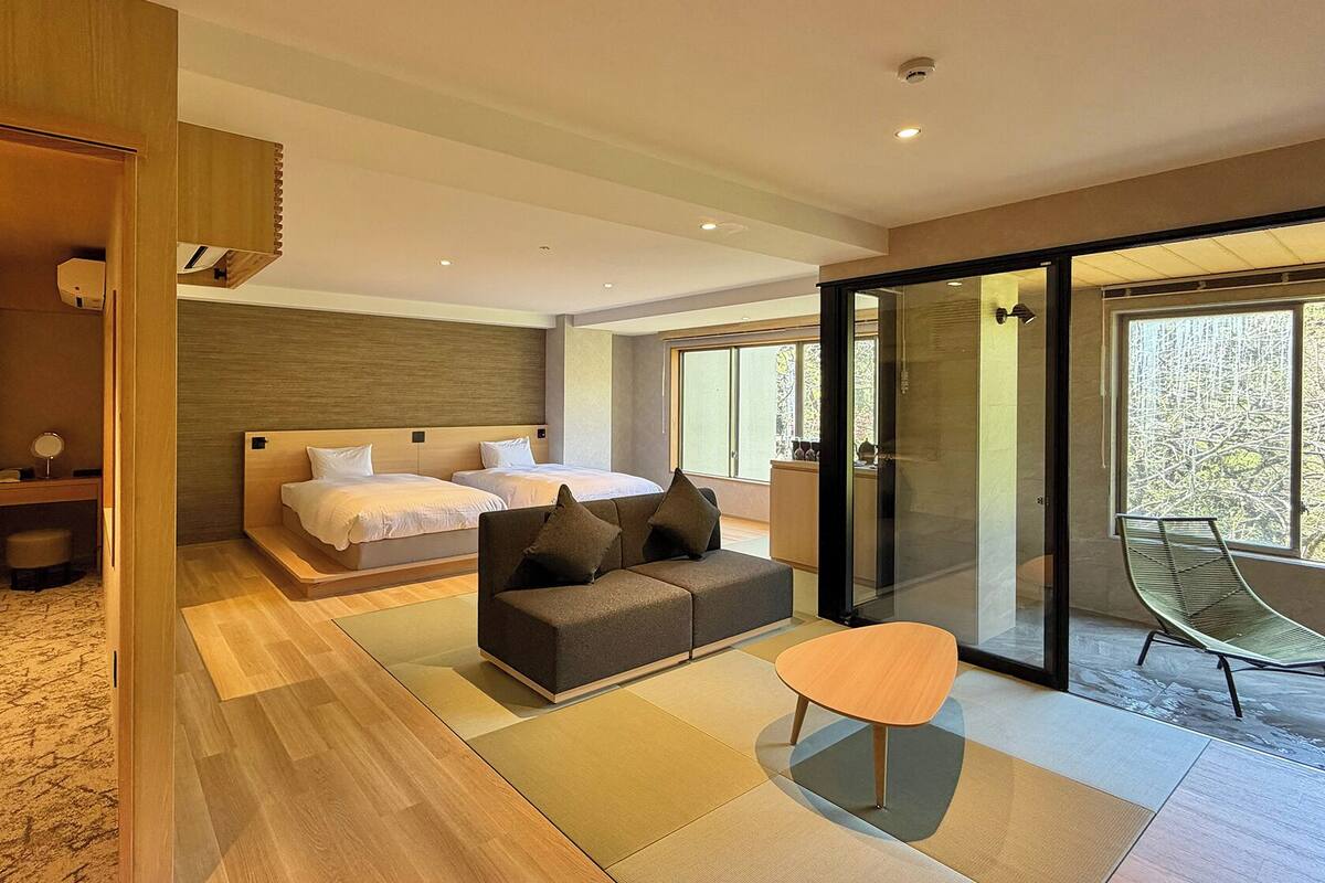 A modern hotel room with a large bed, a sofa, a small table, and a sliding glass door leading to a balcony.