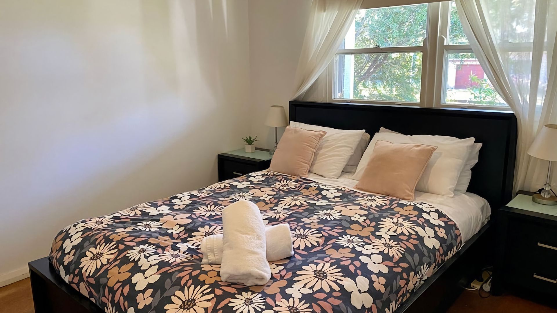 A neatly made bed with a floral bedspread, a bedside table with a lamp, and a window with curtains.