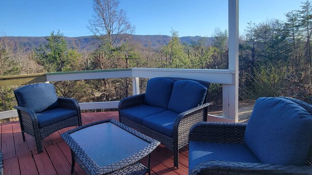 A covered outdoor seating area with wicker furniture and a glass table, overlooking a natural landscape. - Mountain view retreat, 3 bedroom overlooking the Foothills. (Maryville)