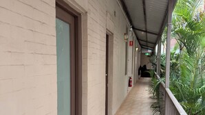 Un couloir couvert doté d’un sol carrelé, d’une rambarde métallique et offrant une vue sur un bâtiment avec une porte et un extincteur.