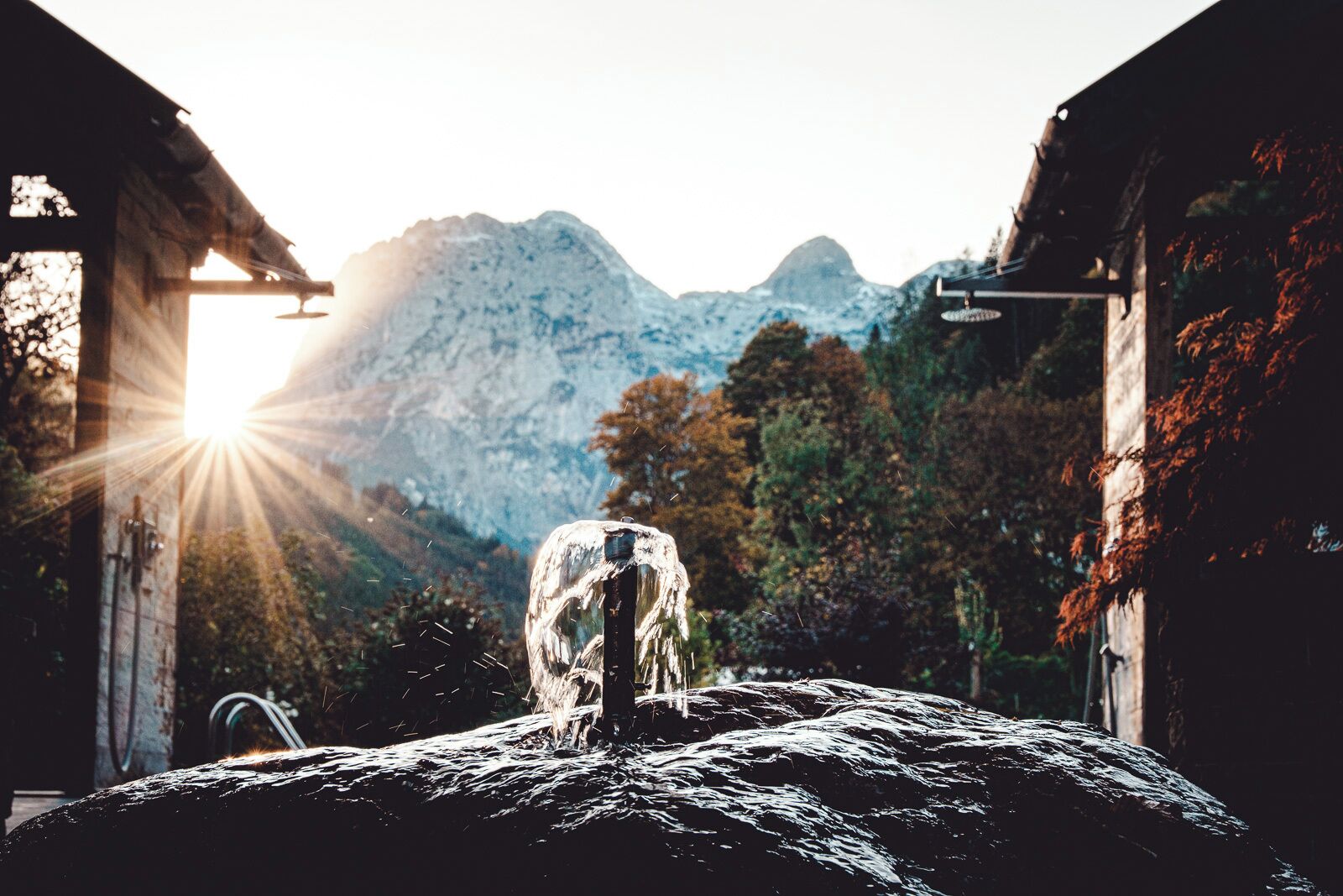 Ein rustikales Steingebäude mit einem Wasserspiel im Vordergrund, vor einer Bergkulisse.
