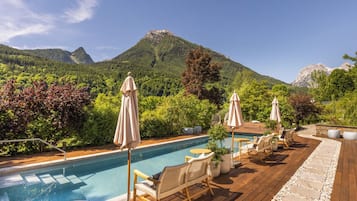 A pool area with lounge chairs and umbrellas, surrounded by lush greenery and mountains.