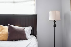 Ein Bett mit weißer Bettwäsche und zwei Kissen, eine Stehlampe und ein Fenster mit Jalousien.