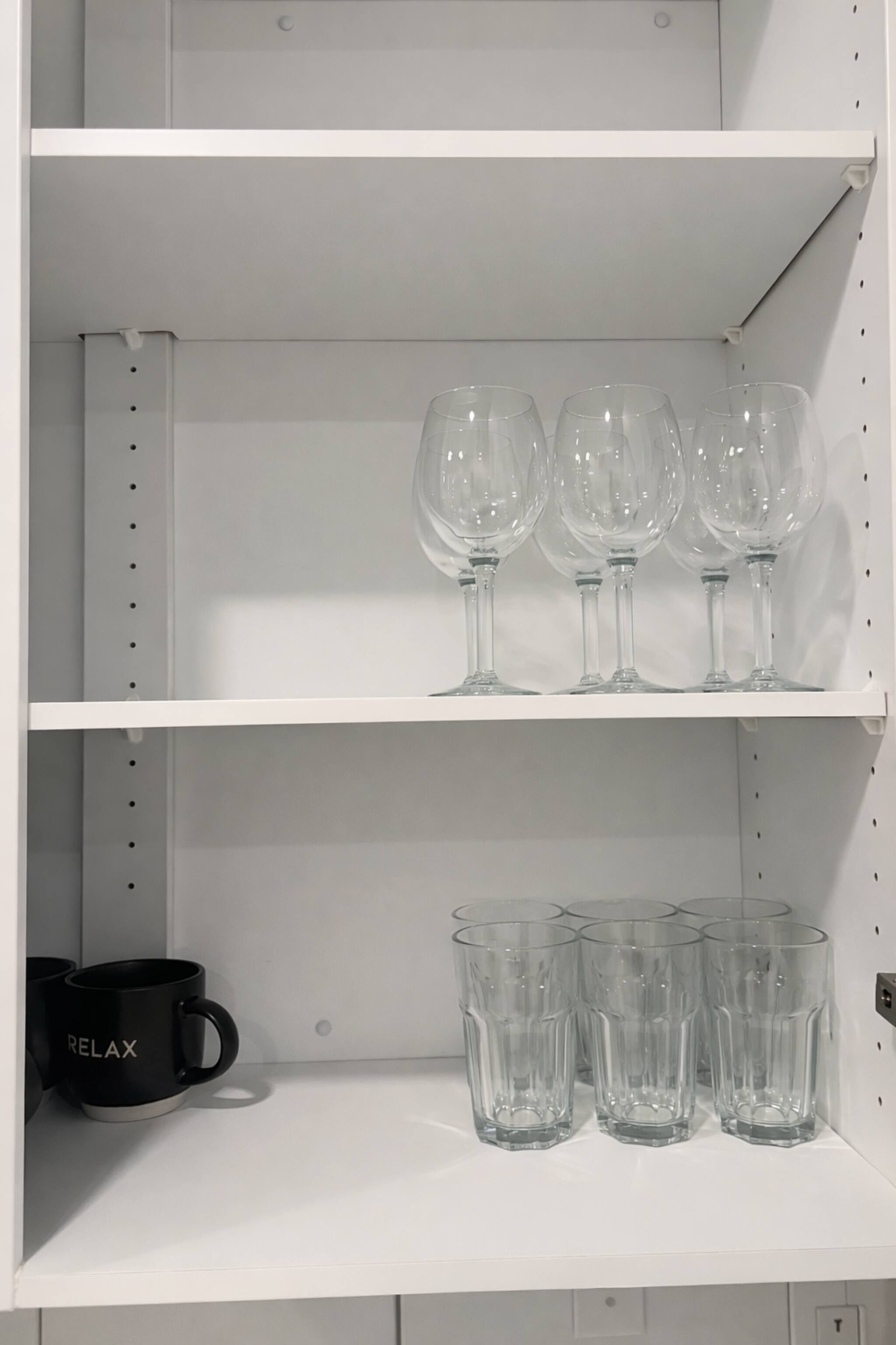 Une étagère blanche avec des verres à vin et une tasse noire sur laquelle est inscrit le mot « RELAX » sur l’étagère inférieure.