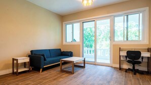 Una sala de estar con un sofá azul, una mesa de centro de madera, una pequeña mesita auxiliar, una silla de oficina negra y un amplio ventanal con vistas a zonas verdes.