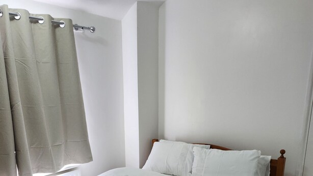 Una cama impecablemente tendida con sábanas blancas y un cabecero de madera en una habitación con paredes blancas y suelo de madera.