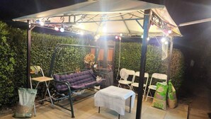 Une terrasse extérieure avec un fauteuil suspendu, une table et des chaises, le tout installé sous une zone abritée et illuminée de guirlandes lumineuses.