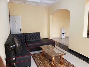 Una sala de estar con un sofá seccional negro, una mesa de centro de vidrio y un ventilador de pie.