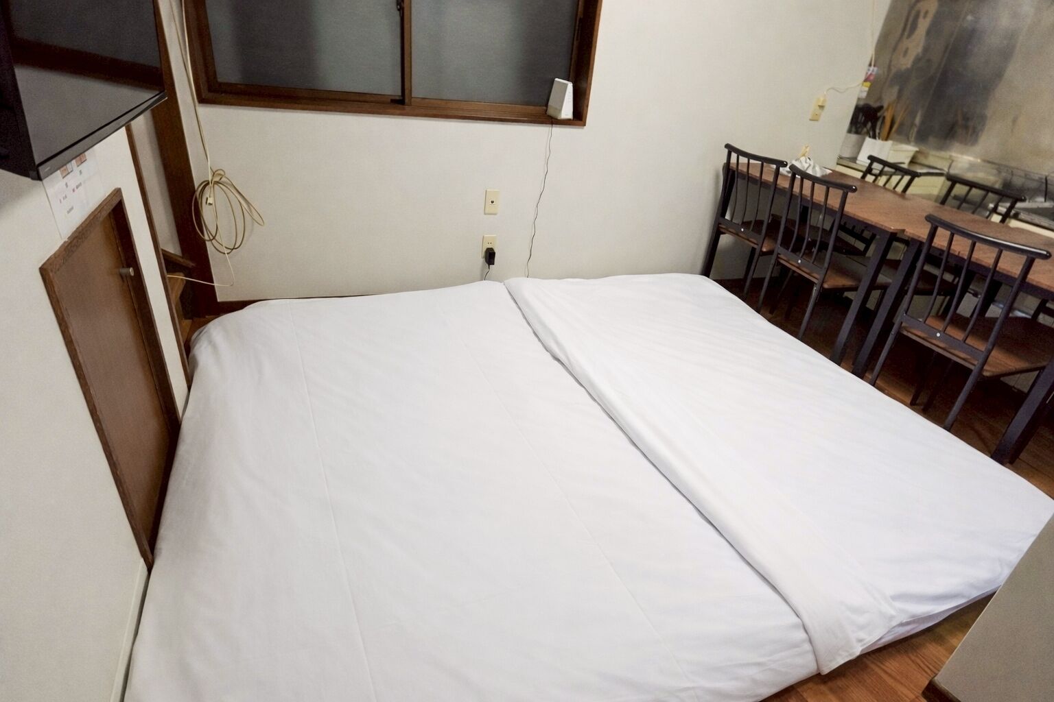 Une petite chambre avec un lit simple, un bureau et des chaises en bois, une fenêtre avec des stores et un haut-parleur fixé au mur.