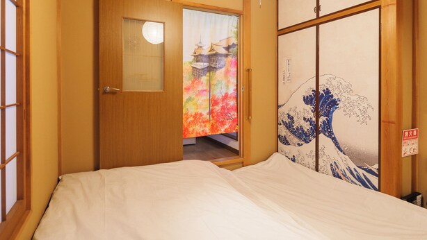 Una habitación japonesa tradicional con techo de madera, cama con ropa de cama blanca, puerta corredera con una obra de arte colorida y un ventanal pequeño.
