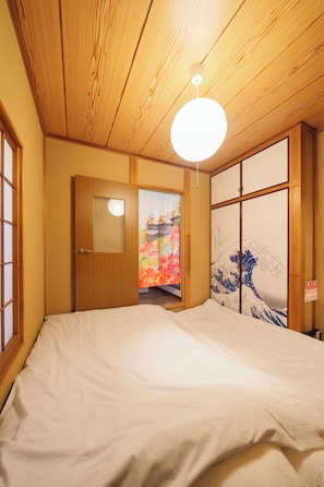 A traditional Japanese room with a wooden ceiling, a bed with white bedding, a sliding door with a colorful artwork, and a small window. - Higashikanagawahouse Yokohama (Yokohama)