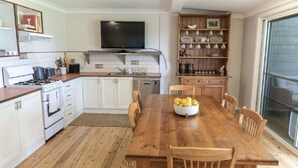 A kitchen with white cabinets, a wooden table with chairs, a TV above the stove, and a shelf with dishes. - Saltwater ~ Cozy sanctuary by the sea (Bendalong)