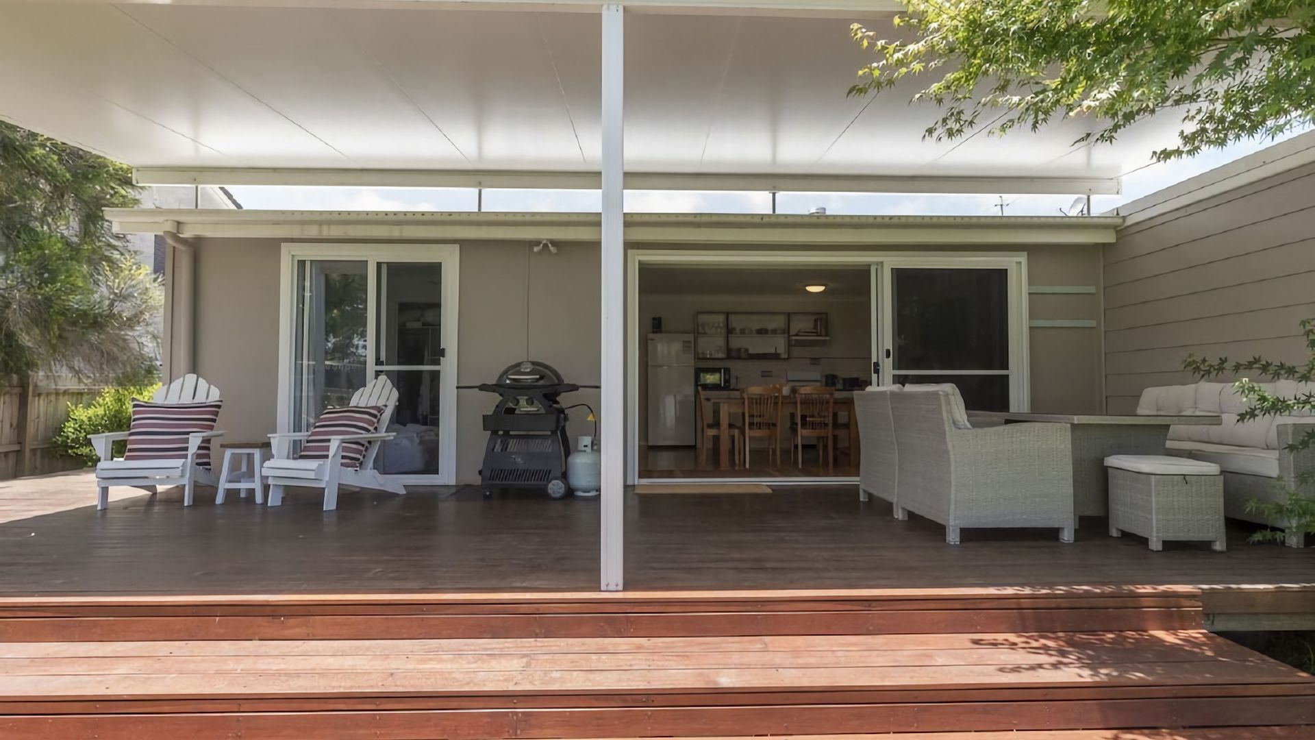 A covered outdoor patio with a wooden deck, white furniture, and a sliding glass door leading to an interior space.