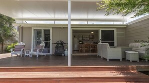 A covered outdoor patio with a wooden deck, white furniture, and a sliding glass door leading to an interior space. - Saltwater is a pet friendly home in Bendalong (Bendalong)