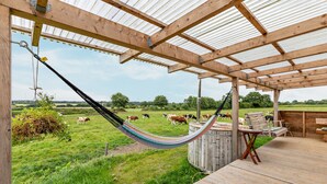 Een houten terras met een hangmat, een houten bankje en uitzicht op koeien die in een weiland grazen.