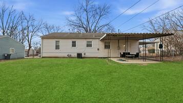 A single-story house with a covered patio area, a lawn, and a tree in the background.