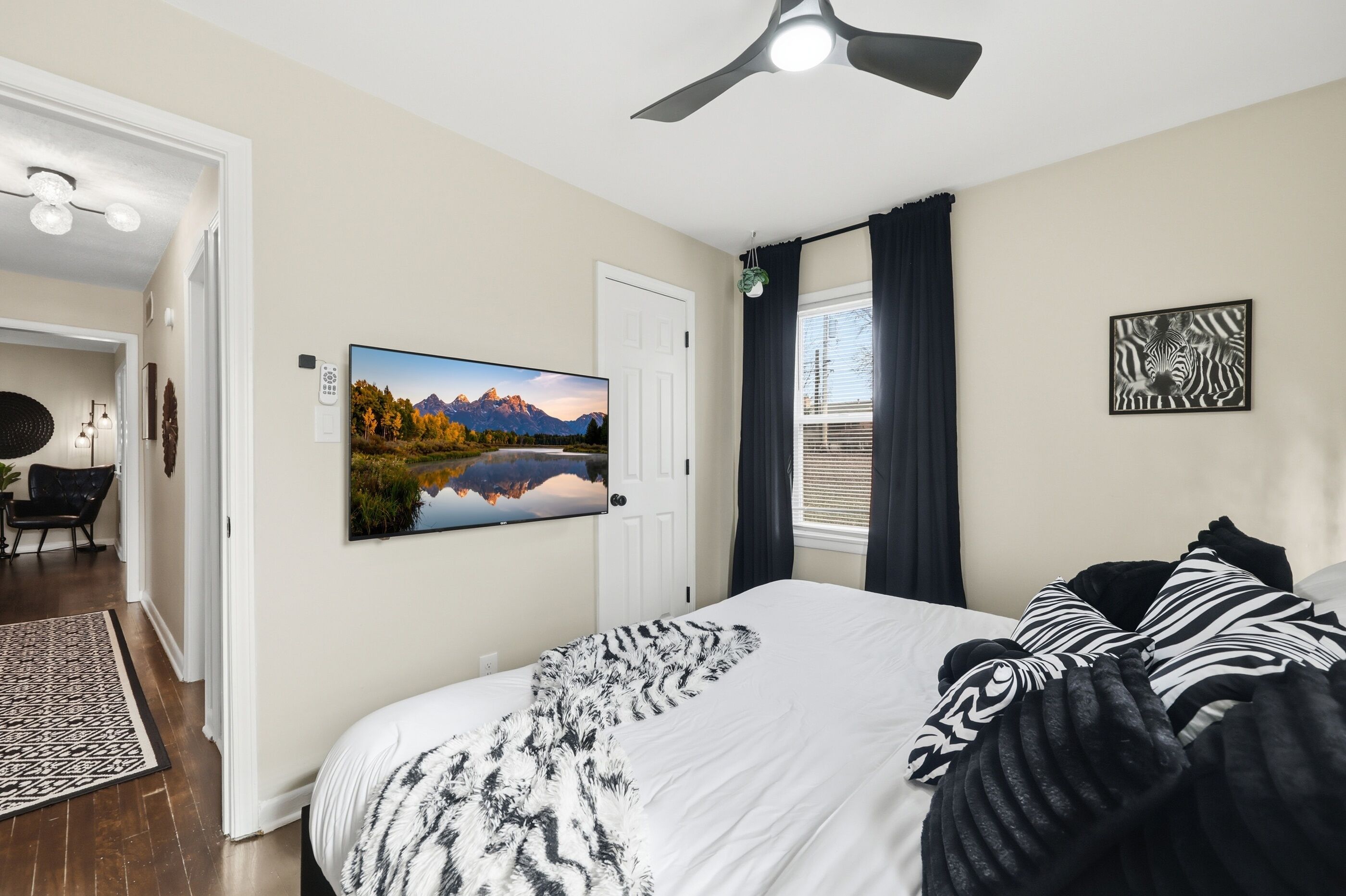 A bedroom with a bed, a painting of a mountain landscape, a ceiling fan, and a window with curtains.