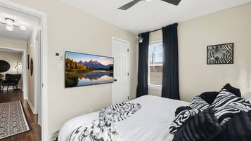 A bedroom with a bed, a painting of a mountain landscape, a ceiling fan, and a window with curtains.