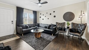 Un salon moderne comprenant un canapé d’angle, deux fauteuils en cuir, une table basse, un ventilateur de plafond, des œuvres d’art décoratives accrochées au mur et une plante en pot.